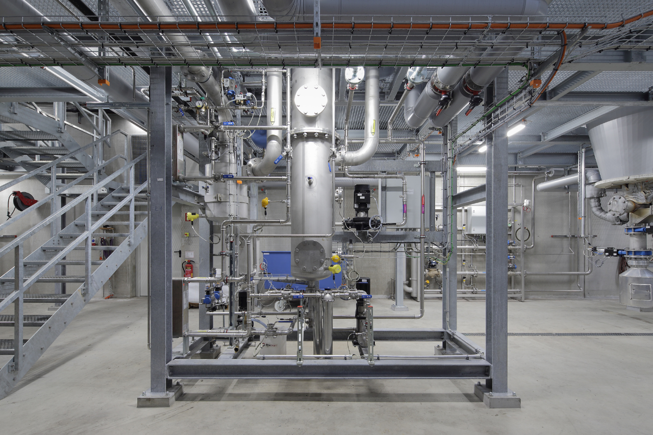 Industrieller Maschinenraum mit Edelstahlrohren, Ventilen, Pumpen und Messgeräten; zentrale vertikale Anlage, Stahlkonstruktion und Treppe links.