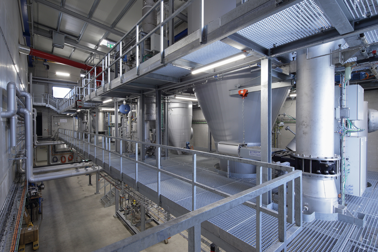 Industrieller Maschinenraum mit Metallkonstruktionen, Treppen, Geländern, Rohrleitungen, großen Silobehältern und technischen Anlagen.