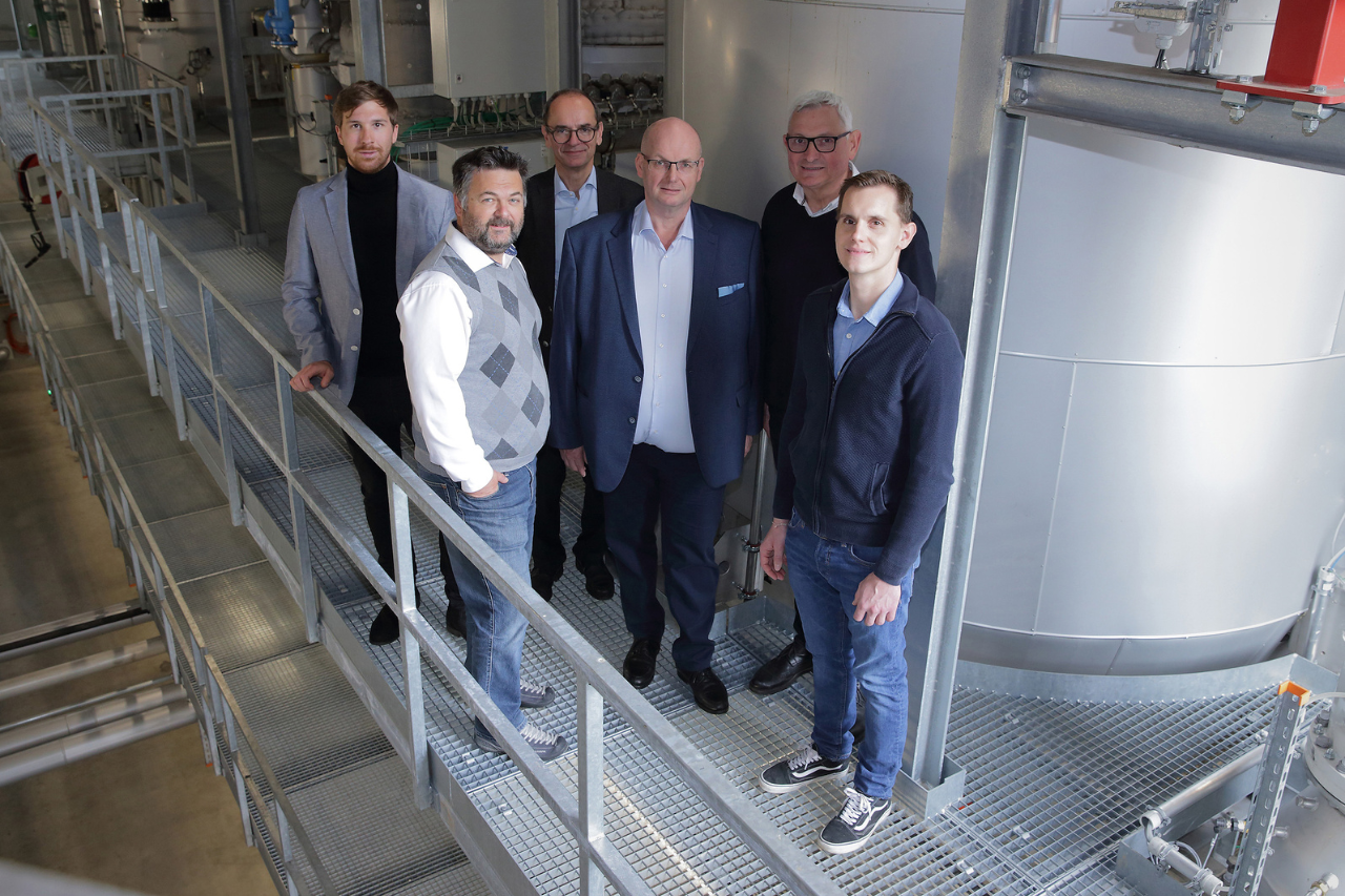 Sechs Personen in Business-Casual-Kleidung stehen auf einer Metallgitterplattform in einer Industrieanlage mit großen Tanks.