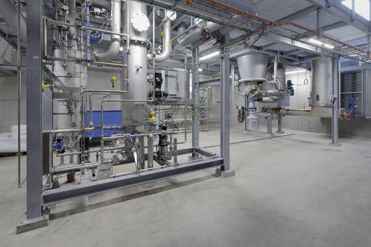 Industrielle Prozessanlage mit Rohren, Ventilen und Tanks in einem Stahlgerüst, Betonboden.