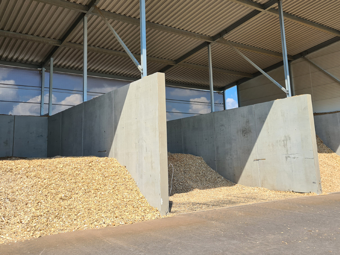 Innenbereich einer Lagerhalle mit Beton-Trennwänden, Holzspäne-Haufen, Stahlkonstruktion und Dach, Himmel sichtbar.