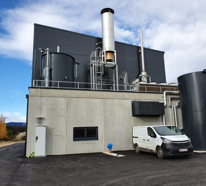 Industrieanlage mit Pufferspeicher, Rohrleitungen und Schornstein; Lieferwagen vor grauer Betonfassade, blauer Himmel.