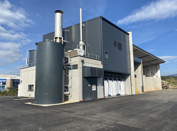 Industrieanlage mit großen zylindrischem Pufferspeicher, grauem Gebäudekomplex, Rohren, Lüftungsschächten und Ladebereich.