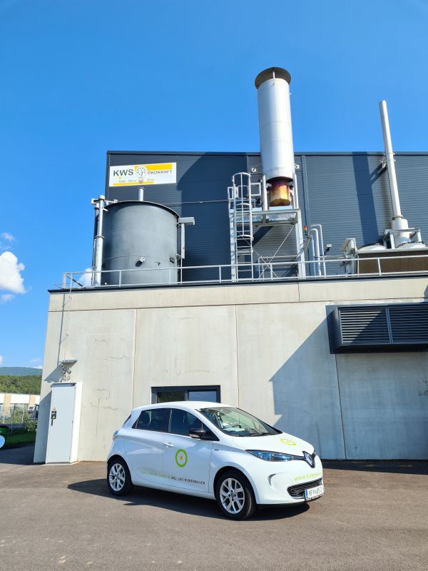 Weißes Elektroauto mit grünem Logo vor einem Industriegebäude mit Turm und Rohrinstallationen, blauer Himmel.
