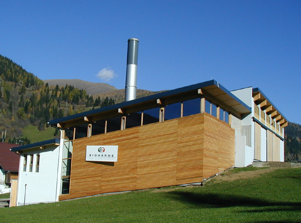 Modernes Gebäude mit Holzfassade, Glasfronten und Schornstein auf grünem Gelände vor Berglandschaft; Schild mit Logo an der Fassade.