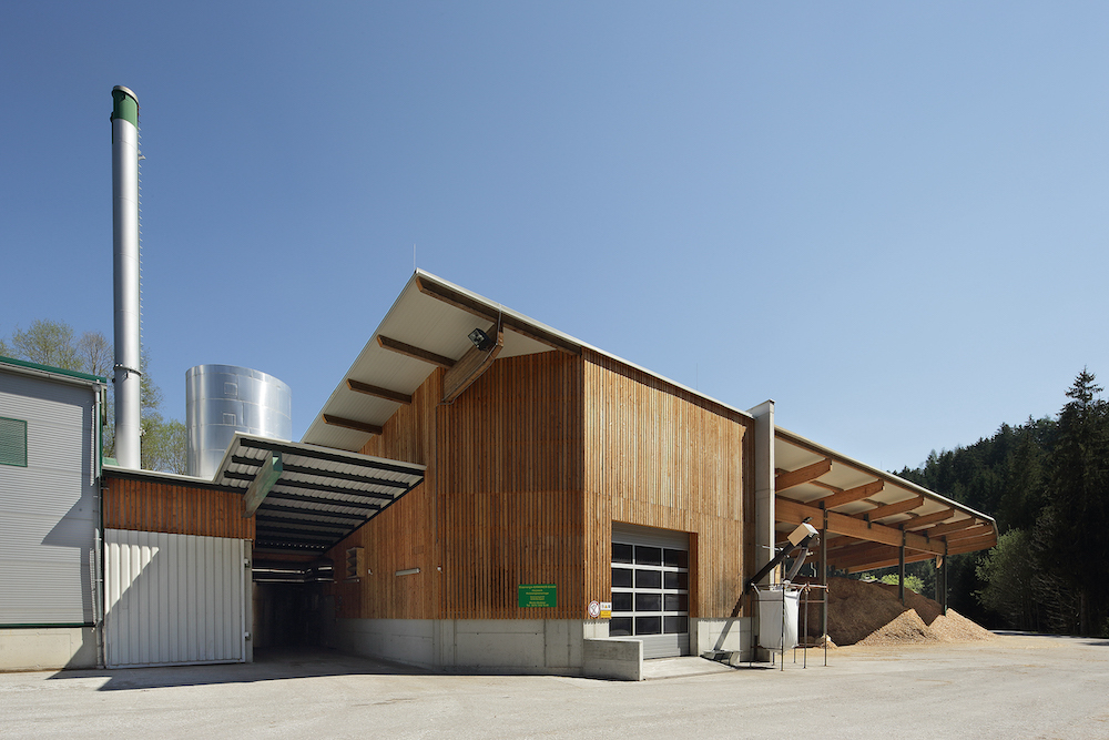 Industrielles Holzgebäude mit Silo, Schornstein und Überdachung; Sandhaufen rechts, blauer Himmel.