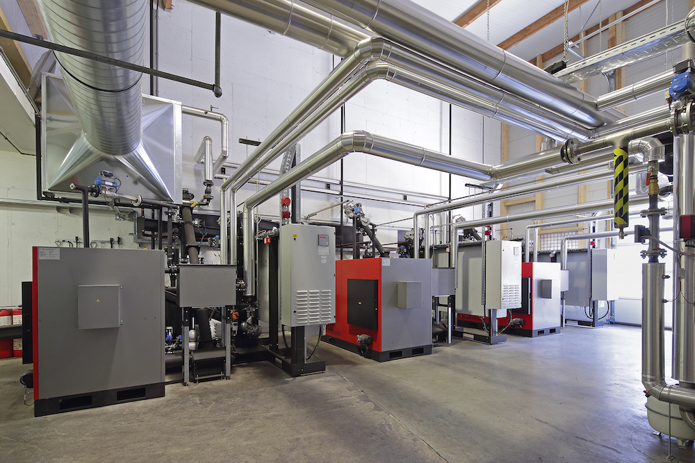 Industrieanlage mit mehreren großen silbernen Rohrleitungen, Kesseln und Steuerkästen in einer Werkshalle.