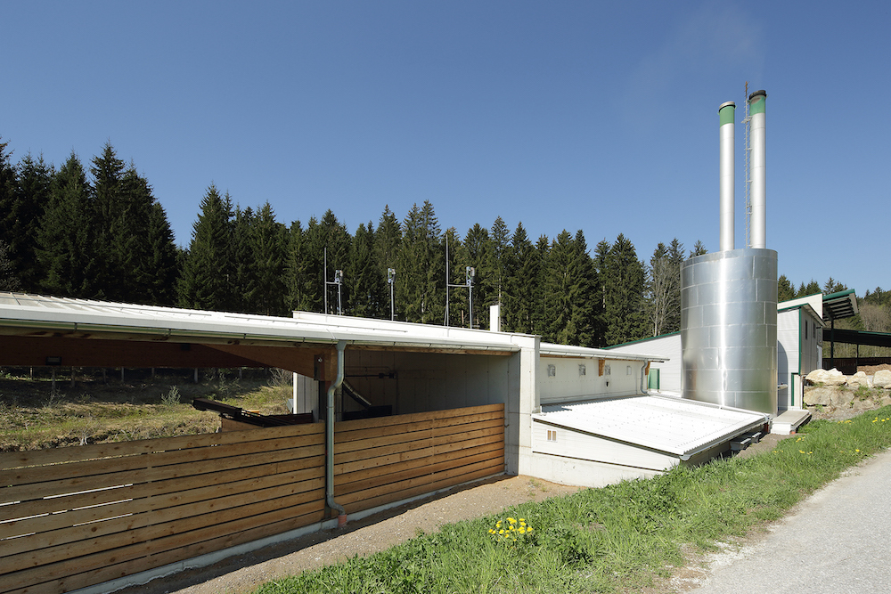 Industrieanlage mit silbernem Pufferspeicher und zwei Schornsteinen, offener Holzunterstand, Wald und blauer Himmel.
