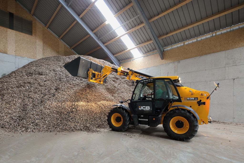 Gelber JCB-Radlader mit Schaufel hebt Holzspäne aus einem großen Stapel in einer Lagerhalle.