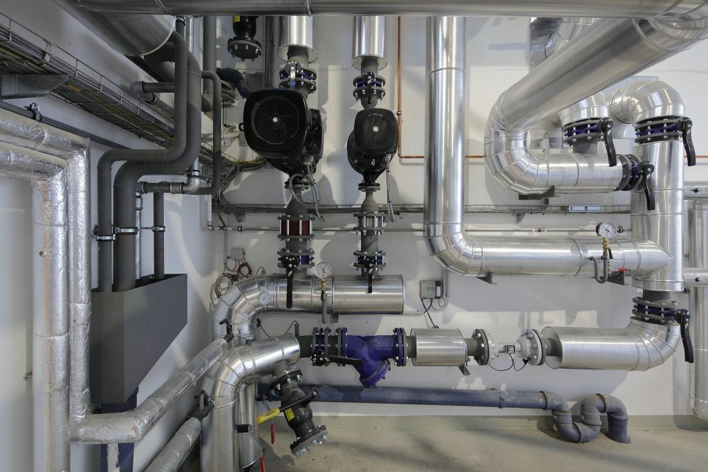 Technikraum mit Metallrohren, Ventilen, Pumpen, Messgeräten und Anzeigen – komplexe industrielle Rohrleitungsanlage.