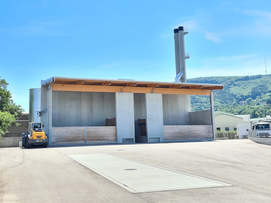 Industrielle Lagerhalle mit Holzvordach, Metallwänden, mehreren hohen Schornsteinen und gelbem Gabelstapler links.