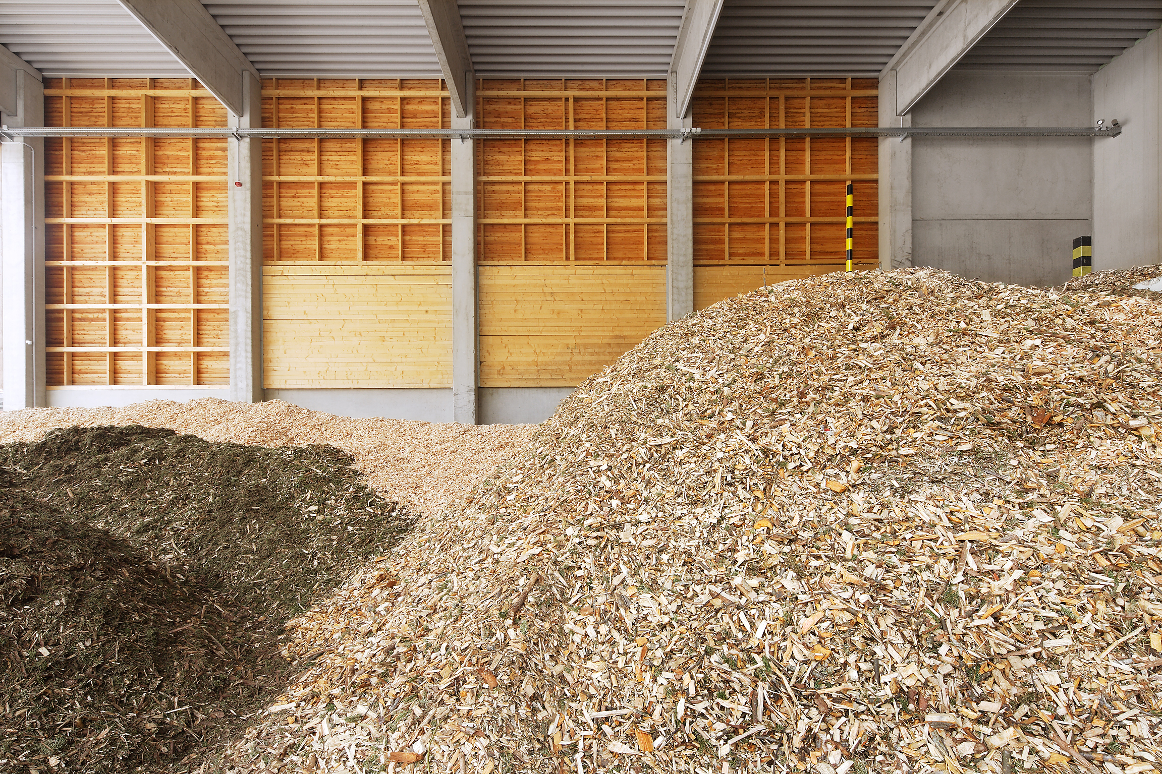 Innenansicht einer Lagerhalle mit großen Holzspänehaufen, Betonstützen, Holzwänden und Deckenbalken.