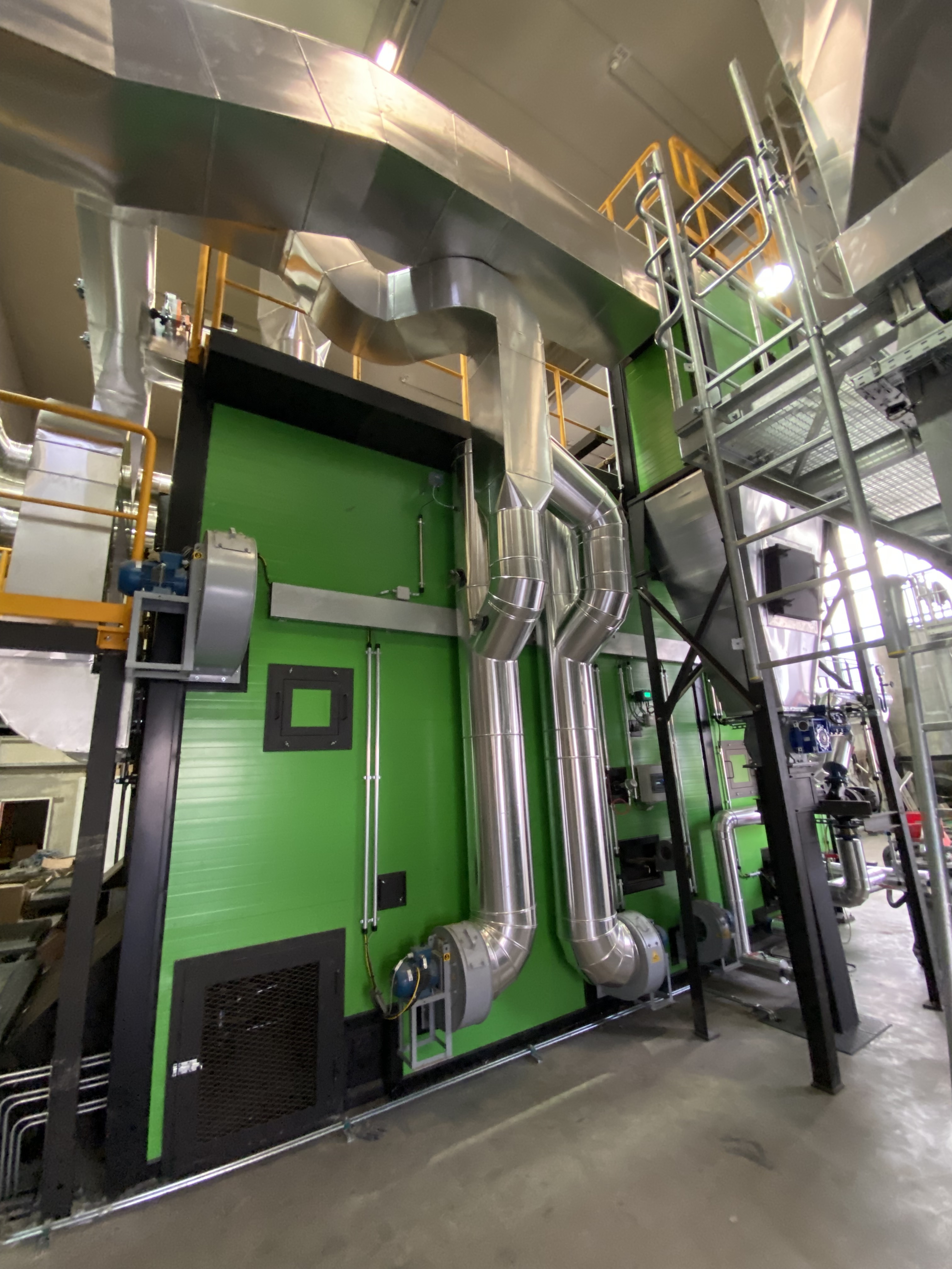 Industrielle Anlage mit grüner Wand, großen silbernen Rohrleitungen, Geländern, Treppen und Maschinen in einer Fertigungshalle.