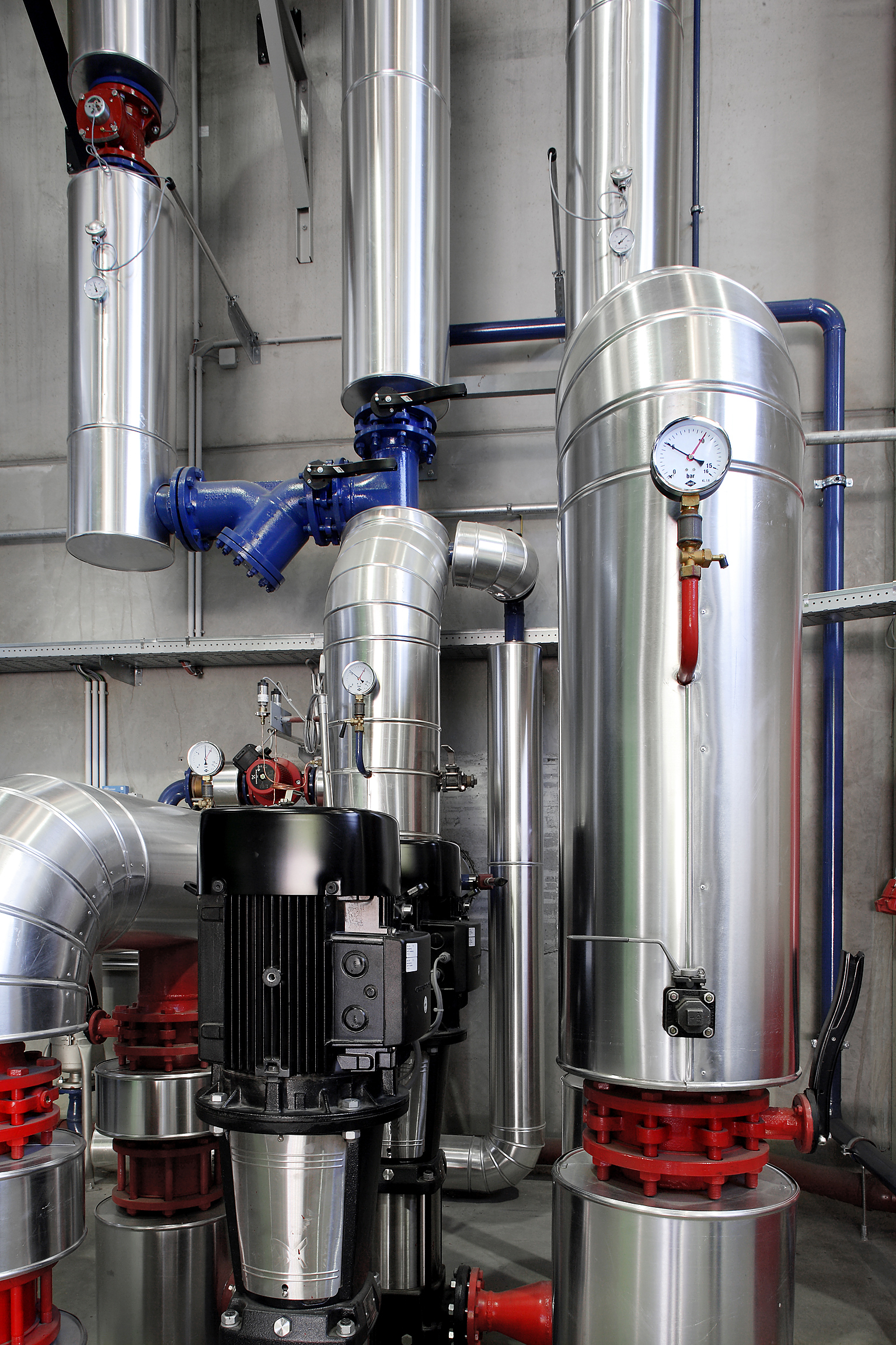 Industrielle Rohrleitungsanlage mit großen silbernen Edelstahlrohren, Messgeräten, blauen Ventilen, Pumpen und rotem Grundrahmen.