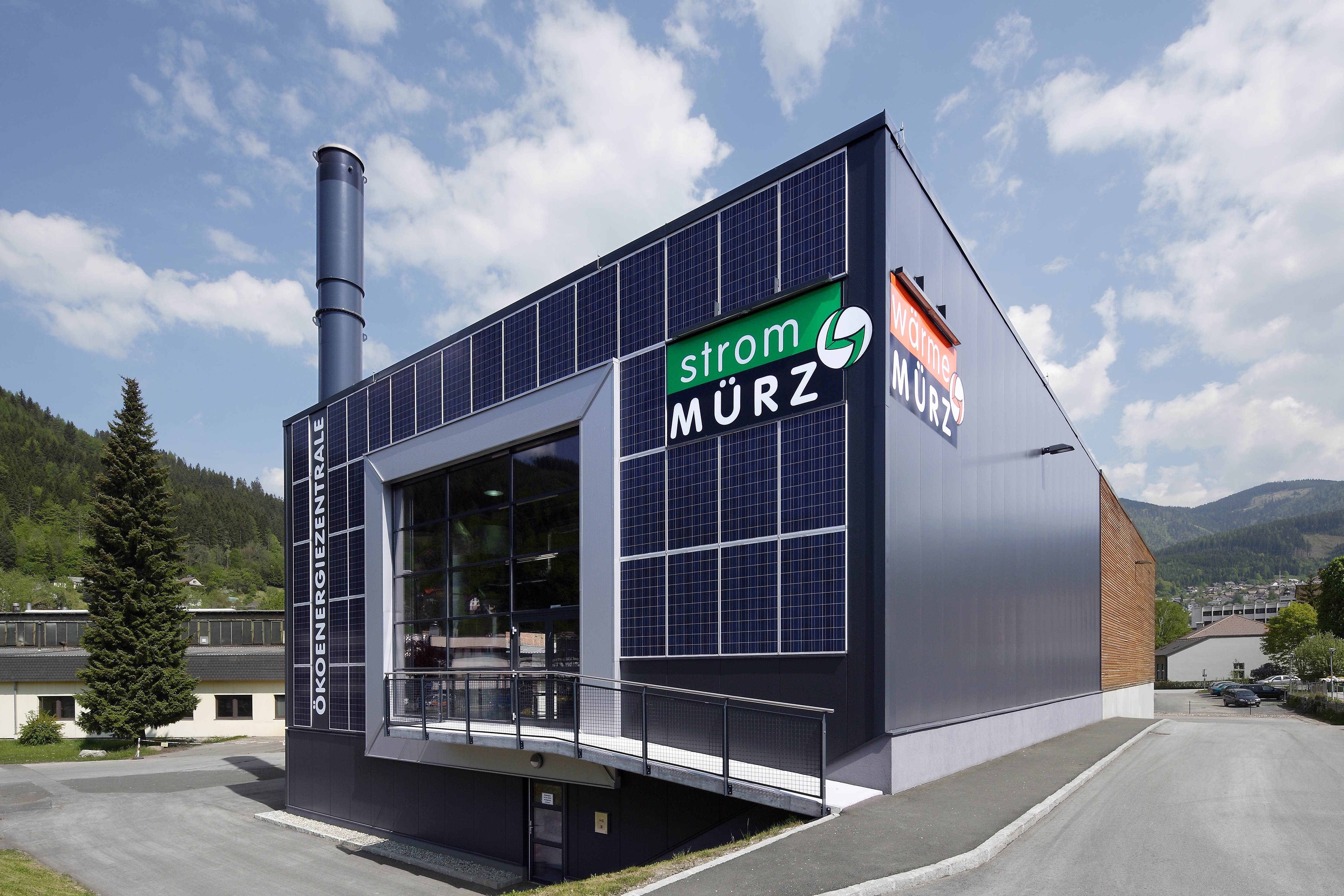 Energiezentrum mit Solarfassade und Glasfront, Schild Strom MÜRZ, Wärme MÜRZ, Schornstein, umliegende Berge unter blauem Himmel.
