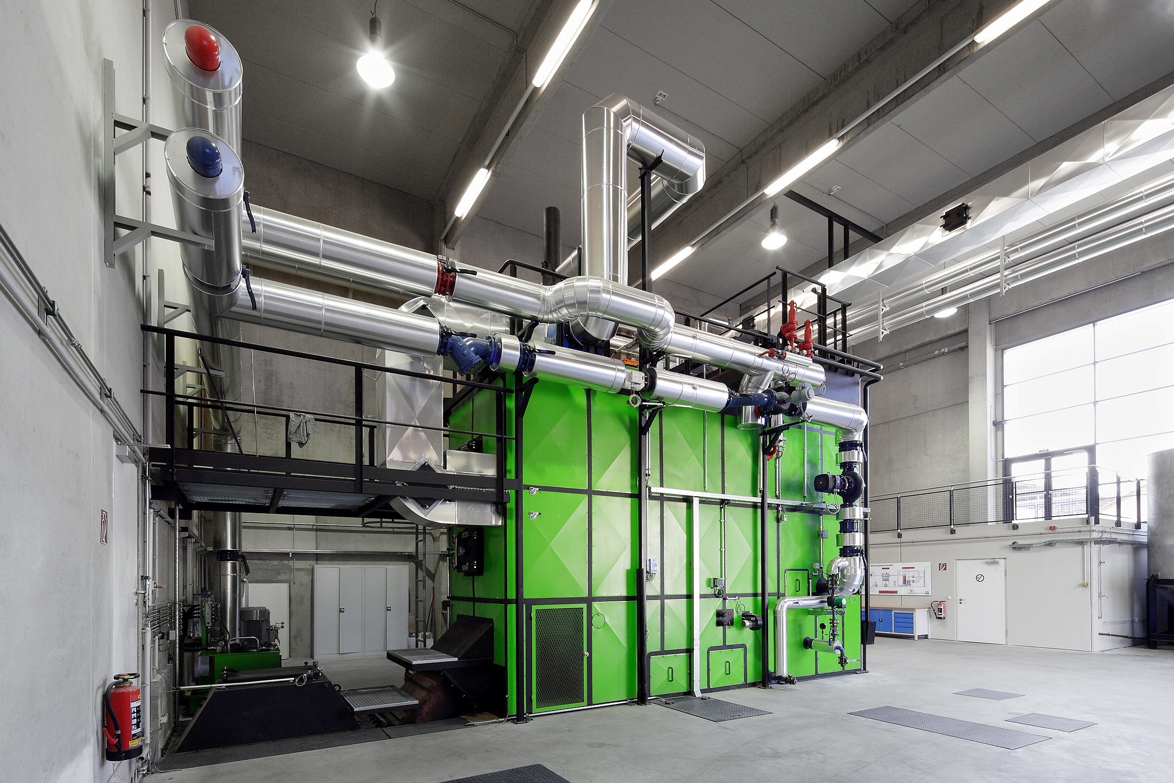 Industrielle Anlage mit grünen Modulen, großen Metallrohrleitungen, Geländerplattform und Deckenbeleuchtung in einer Halle.