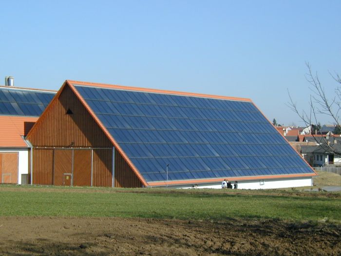 Holzbau mit großem Satteldach, bedeckt von blauen Solarpaneelen; grüne Wiese und Stadt im Hintergrund.