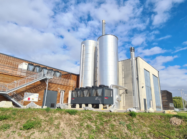 Außenansicht einer Industrieanlage mit zwei silbernen Silos, einem großen Aggregat, Holz- und Betongebäuden, Metalltreppe, unter blauem Himmel.
