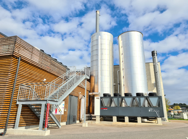 Industrieanlage mit zwei großen silbernen Silos, Metalltreppe, Holzfassade und blauem Himmel.