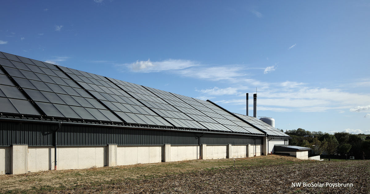 Langgestrecktes Gewerbegebäude mit Solarpanel-Dach, davor Feld; blauer Himmel, im Hintergrund Industrie-Schornsteine, NW BioSolar Poysbrunn