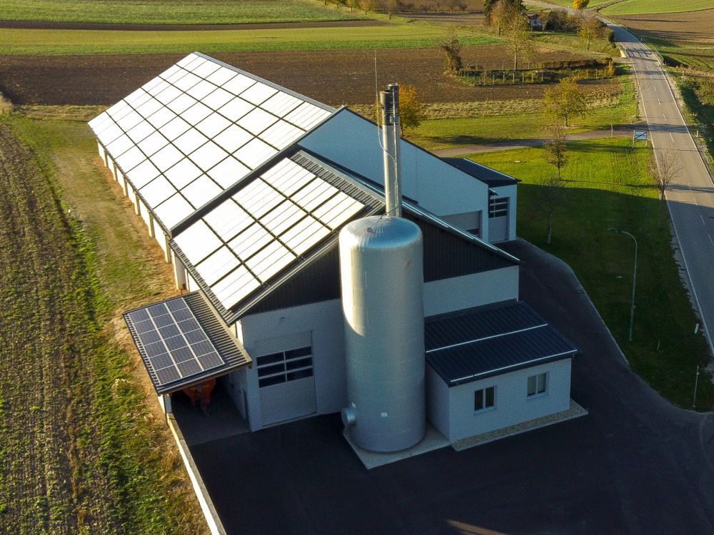 Luftaufnahme eines Industriegebäudes mit Solarmodulen auf dem Dach, einem großen Pufferspeicher und umliegenden Feldern sowie einer Straße.