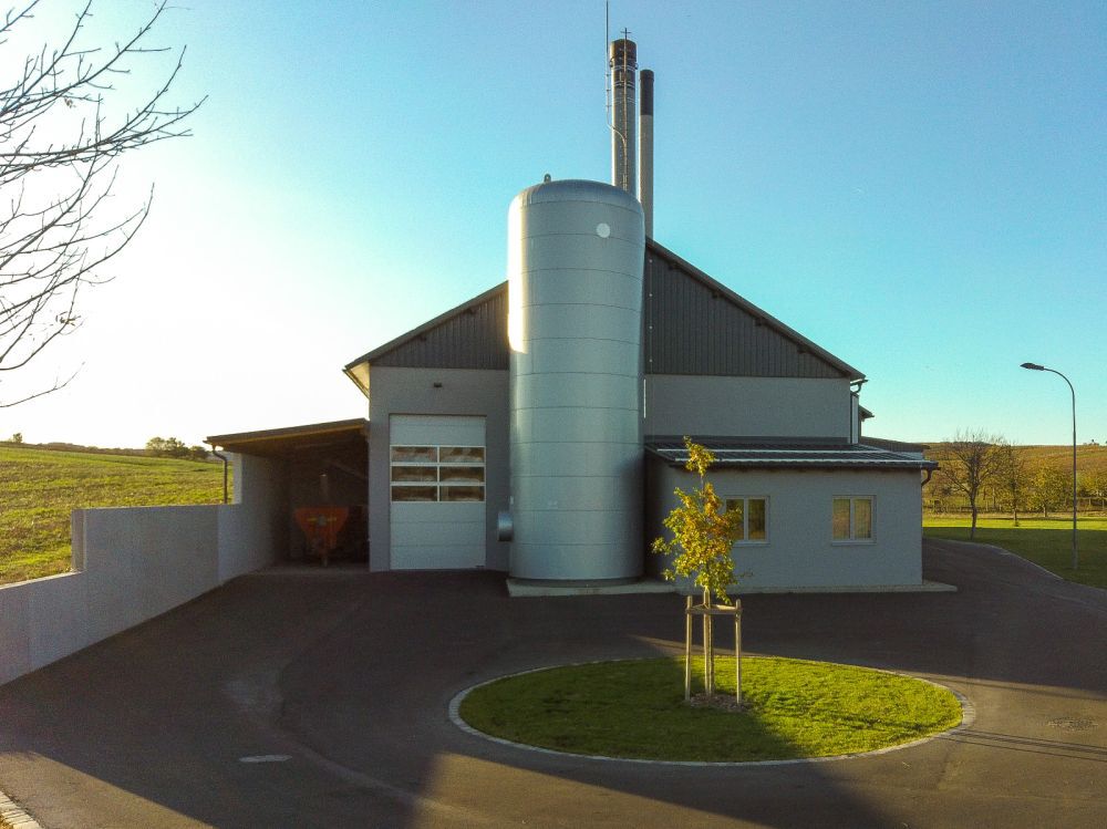Moderne Industrieanlage mit silbernem Pufferspeicher, zwei hohen Rohren, Einfahrt und Kreisverkehr mit Baum, ländliche Umgebung und blauer Himmel