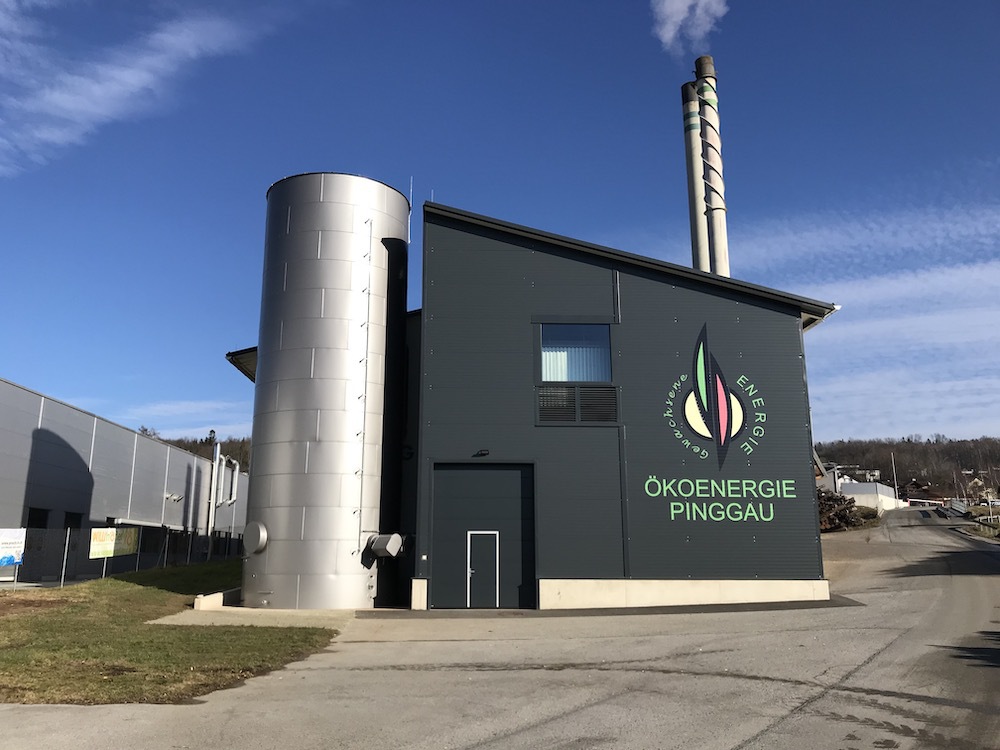 Außenansicht einer Ökoenergieanlage, dunkler Gebäudetrakt, silberner Rundturm, Schlot, Logo ÖKOENERGIE PINGGAU vor blauem Himmel.