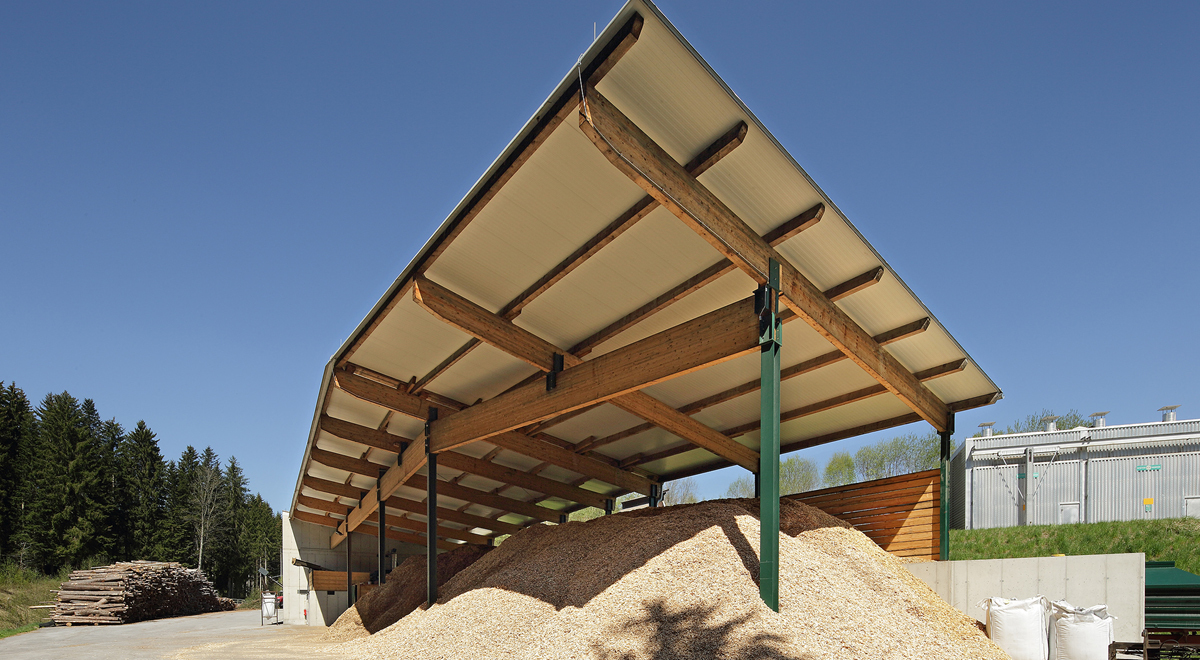 Offene Holzlagerhalle mit Holzbalken und grünen Stützen; großer Haufen Holzspäne und gestapelte Bretter vor Industriegebäuden.