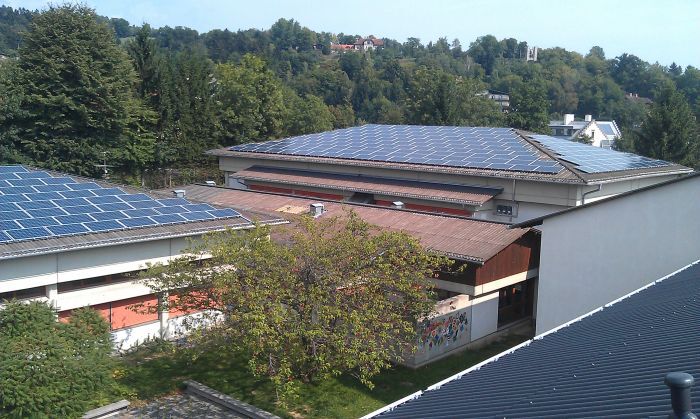 Drohnenaufnahme eines Gebäudekomplexes mit vielen Solaranlagen auf Flachdächern, von Bäumen umgeben.