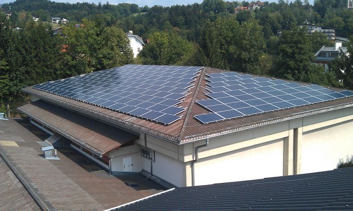 Dach eines Gebäudes mit großer PV-Solaranlage aus Solarmodulen auf dem Flachdach, umliegende Bäume im Hintergrund.