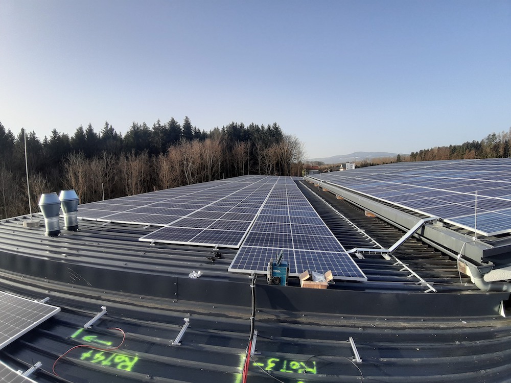 Großflächige Solarzellenanlage auf einem Metalldach; Installationszubehör sichtbar, Wald und blauer Himmel im Hintergrund.