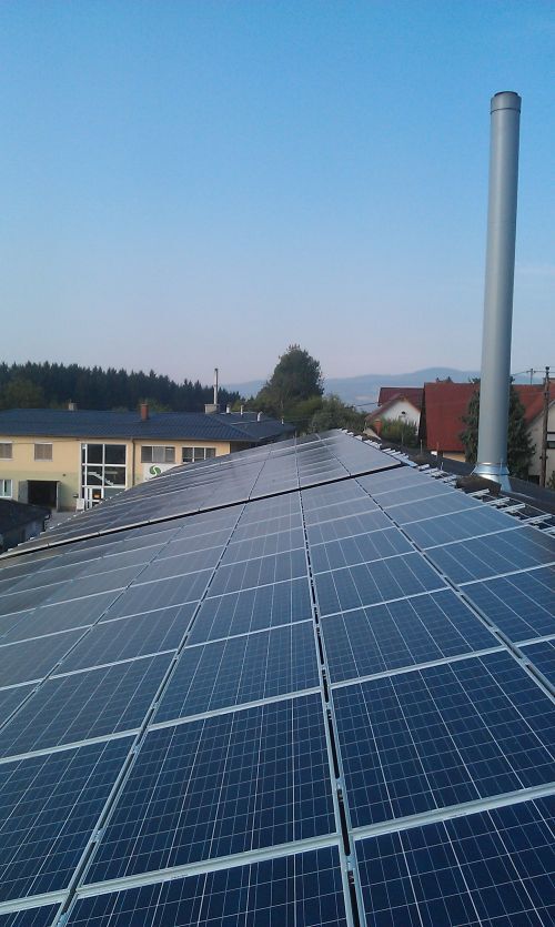 Solaranlagen auf einem geneigten Dach mit vielen Solarzellen; rechts ein Schornstein, im Hintergrund Gebäude, Bäume und blauer Himmel.