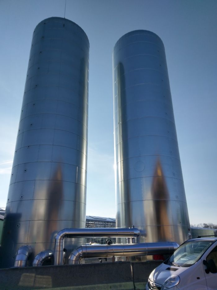 Zwei hohe silberne Pufferspeicher mit Rohrleitungen im Vordergrund; weißer Van am unteren Rand, klarer blauer Himmel.