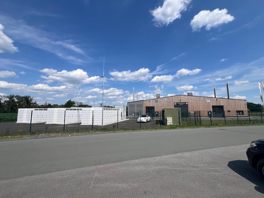 Industrieanlage mit braunem Gebäudekomplex, Zaun, weißen Tanks links, Parkplatz mit Auto, blauem Himmel und Wolken.