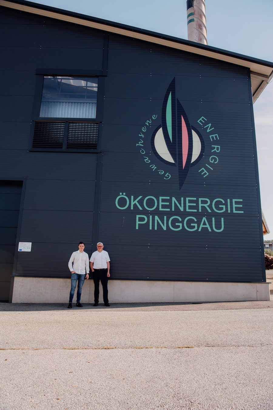 Dunkle Gebäudefassade mit dem Logo ÖKOENERGIE PINGGAU und Schriftzug; zwei neutral gekleidete Personen stehen davor.