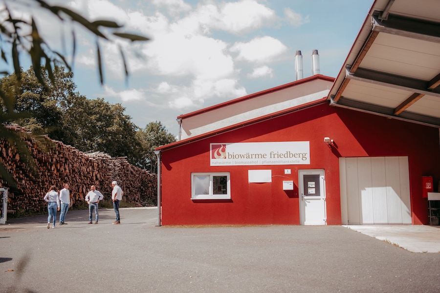 Rotes Gebäude mit Biowärme Friedberg-Signet, vier Personen vor Holzstapeln, Eingangsbereich einer Biomasse-Heizanlage.