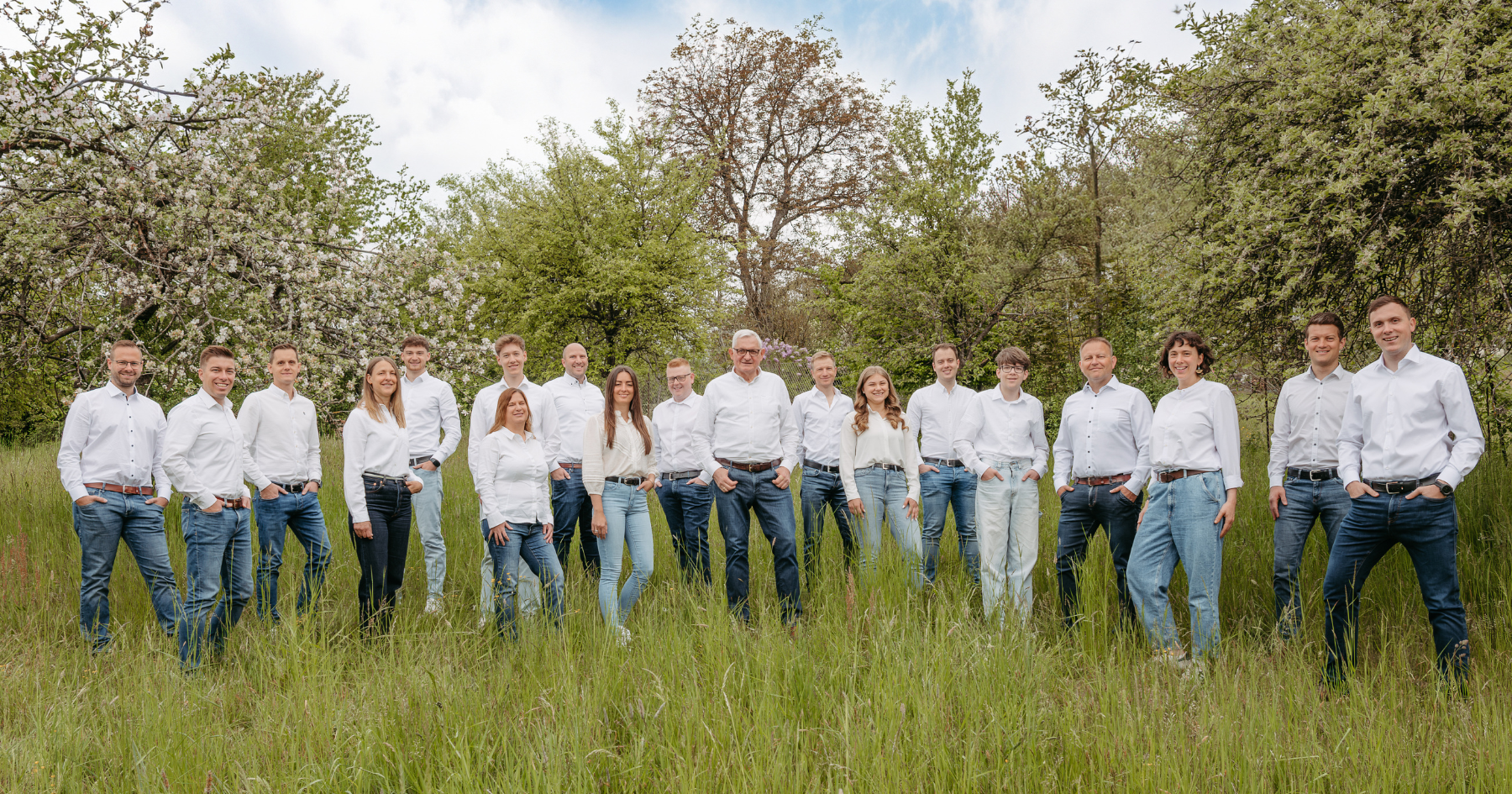 Gruppe von Personen in weißen Oberteilen und Jeans läuft über eine Wiese, Wald im Hintergrund.